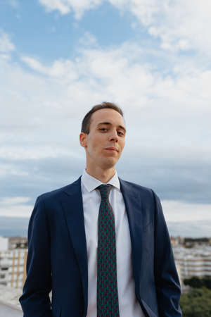 Young businessman in a suit and tie looking at camera and smiling while standing outside in a rooftop with a cloudy skyの写真素材