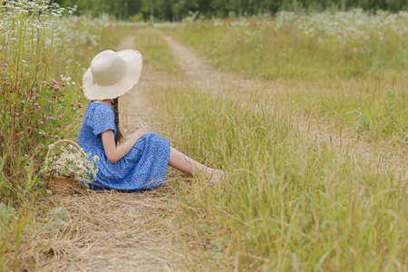 Cute girl in the blue dress on the rural field dreams and looks away towards the horizon