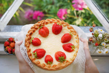 Female hands holding a homemade strawberry cake near window on summer garden background. Fresh...