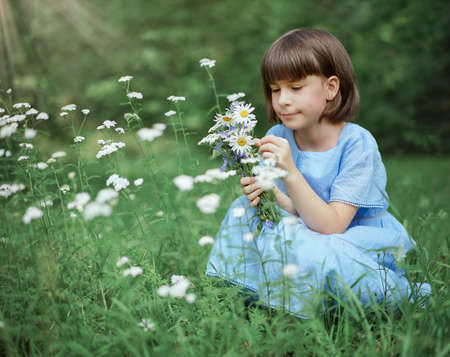 cute little girl in a blue dress with a bouquet of daisies sits on a flowers meadow in the parkの写真素材