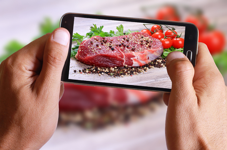 Male hand taking photo of Raw beef steak with spices and cherry tomatoes on wooden background with cell, mobile phone.の写真素材