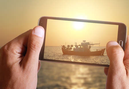 Male hand taking photo of a boat in the sea in sunset with cell, mobile phone.の写真素材