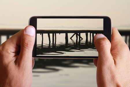 Male hand taking photo of old wooden dock in sunset with cell, mobile phone.の写真素材