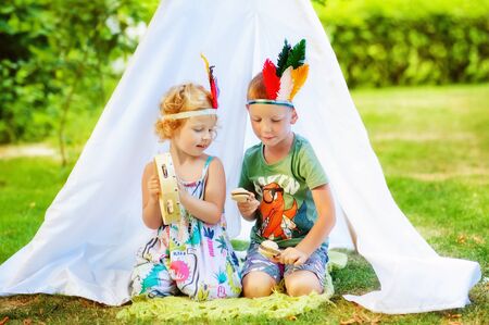 3 years old girl and 5 years old boy with feathers on head sit near white tent with tambourine and maracas in hands. Children pretend to be indian. Indians game in summertime. Children play music.の写真素材