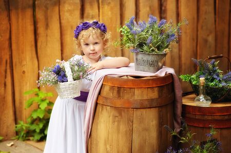 Small girl in white clothes and violet wreath in rustic style decorations. Ecolife. Organic and ecological lifestyle in childhood. Wooden background and flowers around. Child in antique dress.の写真素材