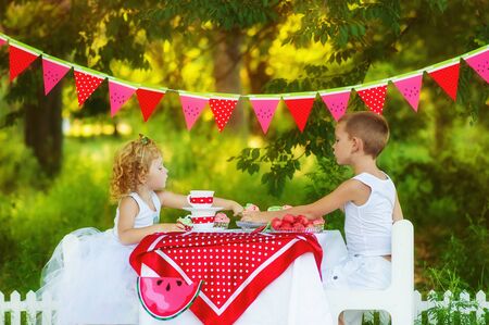 Two children brother and sister eat and drink on beautiful decorated picnic. Hungry kids boy and girl eating outdoors. What to do in summer with children. Watermelon decorated t-shirts.の写真素材