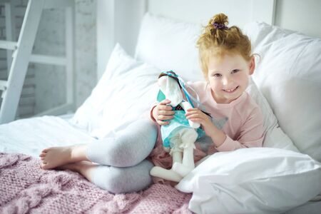 Portrait of little smiling 5 years old girl with curly blond hair laying in pink and gray pajama in her bed with her favorite rabbit toy. Day of sleep. Happy childhood in cozy home with parents careの写真素材