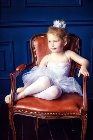 Portrait little 4 years old blonde curly ballerina sitting in dark room on brown leather chair in ballet dress white tutu and pointes. Relaxation in ballet studio. Dancing is hard work.の写真素材