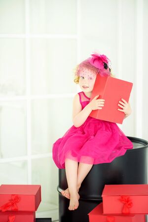 Little 4 years old girl in beautiful pink dress and pink hat with veil sitting on black barrel with red gift box in hands with presents around. Birthday girl waiting for friends on birthday partyの写真素材