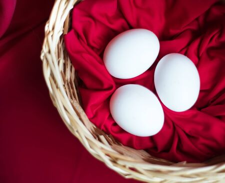 Top view on three white chicken eggs on red fabric in willow brown basket in Easter time. Healthy organic homegrown eating. Easter traditional food. Red easter background. Copy space for textの写真素材