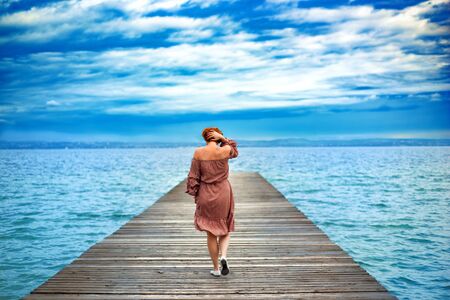 Back of faceless red haired woman in red dress walking by wooden pier on Garda lake in cloudy day. Italian vacation. Garda lake great tourist destination and attraction in North Italyの写真素材