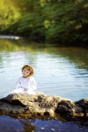 Vertical portrait of 5 years old girl in white traditional embroidery ukrainian dress and chamomiles wreath on rocks in river in Carpathian mountains. Travel tourism destination in Ukraineの写真素材