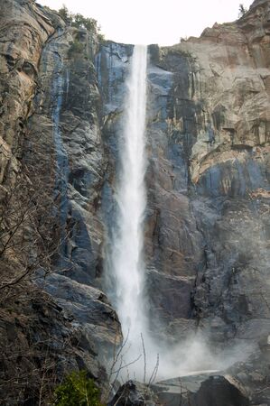 Vertical scenic landscape with waterfall, rocks and large sequoia in Yosemite valley, Yosemite national park, California, USA. Travel tourism hiking destination. View of waterfall in cloudy day.の写真素材
