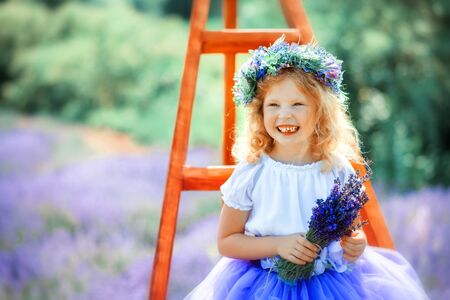 Portrait of happy cute little blonde curly girl wearing lavender dress and wreath and holding bouquet of lavender in lavender field with violet flowers around in sunny summer day. Summer in Provence.の写真素材