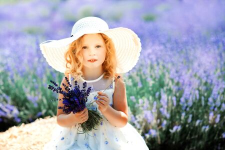 Portrait of happy cute little blonde curly girl wearing white dress and hat and holding bouquet of lavender in lavender field with violet flowers around in sunny summer day. Summer in Provence. Travelの写真素材