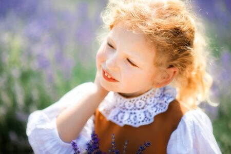 Close-up portrait of cute little tired blonde curly girl wearing vintage brown dress with lace sitting in lavender field in sunny summer day. Summer in Provence. Travel. Card in vintage styleの写真素材