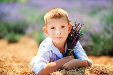 Portrait of cute little blonde boy wearing white shirt sitting on straw in lavender field with bouquet of lavender in sunny summer day. Summer in Provence. Travel. Vintage styleの写真素材