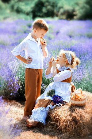 Vertical picture of children in vintage clothes in middle of lavender field. Girl in brown dress with white apron and boy in white hat, shirt and brown trousers have picnic on lavender. Eating outdoorの写真素材