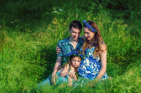 Pregnant woman mother to be in wreath and blue dress sitting with her daughter and husband in high green grass in sunset time in summer. Pregnancy and healthy organic close to nature lifestyleの写真素材