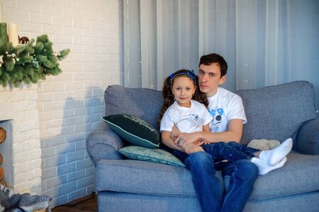 Sweet little girl together with beautiful young dad sitting on sofa in living room at home in evening. Happy family concept. Love. Trust. Togetherness. Parents and children. Family time. Stay home.の写真素材