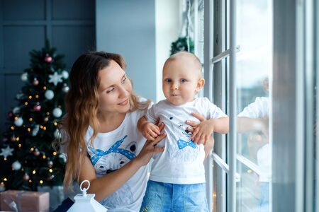Portrait of mother hugging smiling son at home on Christmas eve. Happy young mother with baby against Christmas lights. Happy family mother and toddler near Christmas tree near window in winter.の写真素材