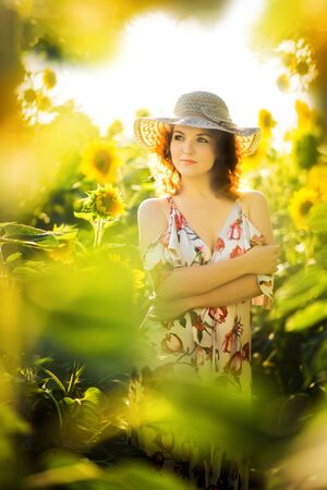 Vertical portrait of young beautiful red haired woman in straw hat in sunflowers field in back light. Summer countryside concept. Close to nature vacation. Woman and sunflower. Summer light. Outdoorsの写真素材