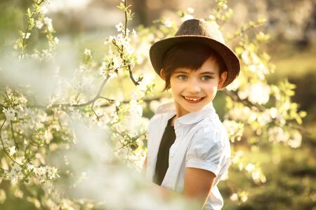 Portrait of 7 years old cute stylish short haired smiling girl in white shirt and brown hat looking away in blooming garden in spring. Short haircut for girl. Allergy seasonの写真素材