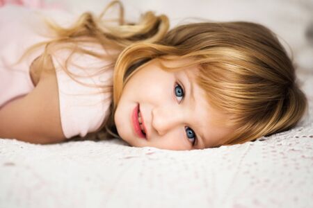 Close-up portrait of beautiful cute little 3 years old blond girl with blue eyes lying on bed in pink dress. Moment of relaxation for active child. Kid is bored indoorsの写真素材