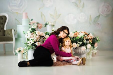 Young beautiful brunette mother in glasses hugging little 3 years old blonde girl in pink dress. Portrait of woman, child and white rabbit. Care, love, trust, togetherness. Family time in Easter.の写真素材