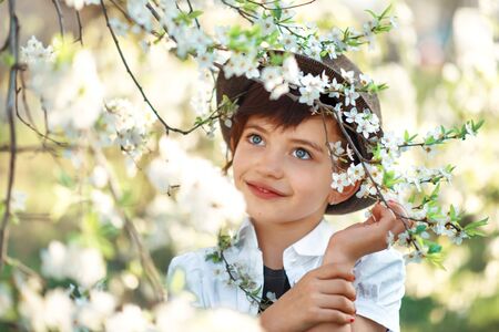 Portrait of 7 years old cute stylish short haired smiling girl in white shirt and brown hat touching her hand in blooming garden in spring. Short haircut for girl. Allergy season. Outdoorsの写真素材