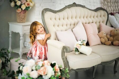 Happy smiling blonde 3 years old girl playing hide and seek in bright living room with sofa and bouquets of flowers everywhere. Indoors spring photoshoot of small joyful child. Games in self isolationの写真素材