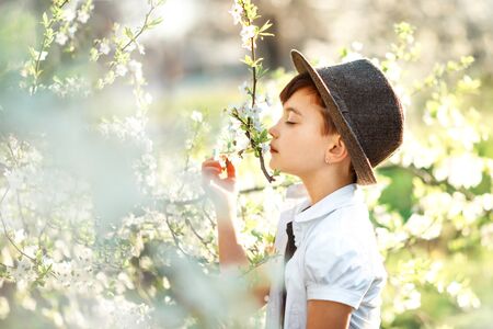 Portrait of 7 years old cute stylish short haired smiling girl in white shirt and brown hat sniffing flower in blooming garden in spring. Short haircut for girl. Allergy season. Non allergic child.の写真素材
