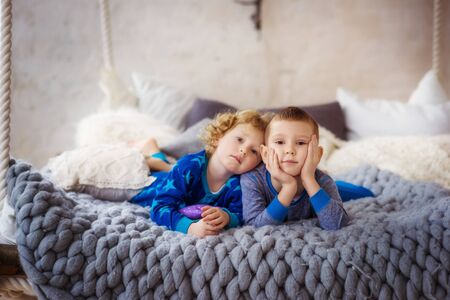 Kids laying in bed with gray cover in white bedroom. Little boy and girl, brother and sister, siblings play on the bed wearing matching pajamas. Family at home. Bored children in self isolationの写真素材
