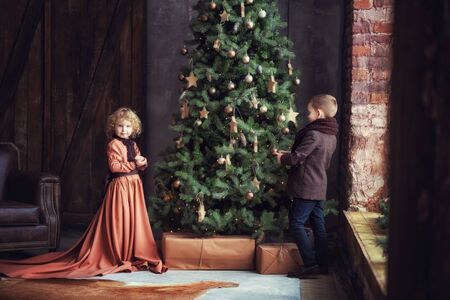 Two children boy and girl brother and sister siblings in brown vintage retro clothes decorating big christmas tree in dark room near window. Curly blonde girl and her brother. Happy new year and xmas.の写真素材