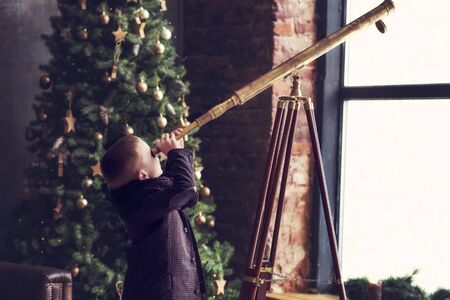 6 years old boy in brown vintage clothes looking in telescope with christmas tree on background. Little boy explore world. Retro post card with children. Child holding telescope. Self educationの写真素材