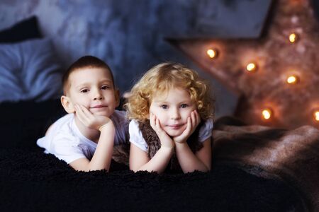 Two children, boy and girl, brother and sister, siblings, laying in bed in loft styled bedroom with star on background. Curly blonde girl and brother. Kids spend time together. Family. Self Isolation.の写真素材