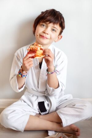 Vertical portrait of cute 7 years old hungry short haired girl in white kimono eating pizza after hard on-line karate taekwondo training. Child after distance training eat at home. Martial arts.の写真素材