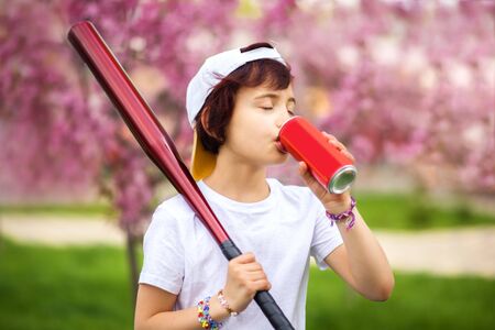Happy caucasian little girl in white turned back cap with soda refreshment in can and baseball beat in park in spring time with blooming tree around. Child outdoors. Nutrition, sport, activityの写真素材
