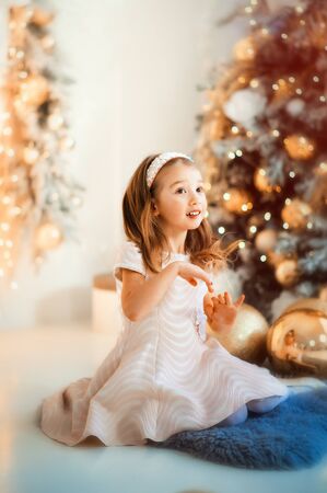 Vertical portrait of small 4 years old beautiful surprised girl in white fashion dress sitting on floor near christmas tree with christmas light around. Xmas and new years eve concept. Happy holidaysの写真素材