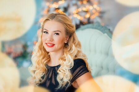 Beautiful long haired blonde young woman in black fashion dress sitting on sofa near christmas tree with christmas light around. Xmas and new years eve concept. Happy holidaysの写真素材