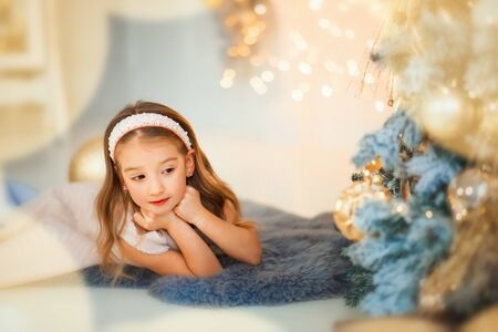 Small surprised 4 years old beautiful girl in white fashion dress laying on floor and waiting for presents near christmas tree with christmas light around. Xmas and new years eve. Happy holidaysの写真素材