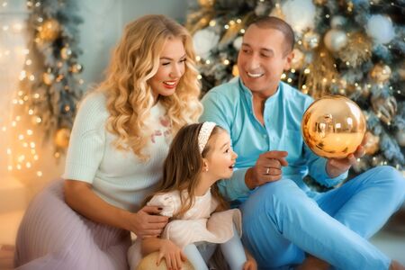 Family Christmas. Lovely family in bright clothes play near decorated Xmas tree on winter evening. Happy household with daughter on floor at home. Merry Christmas, happy New year concept. Trust, love.の写真素材