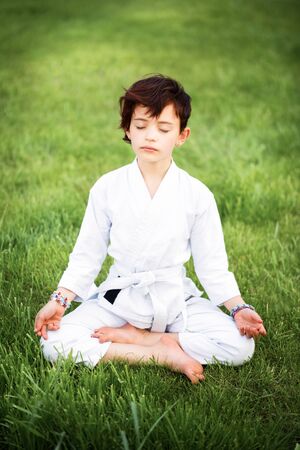Vertical picture of short haired 7 years old girl in white kimono during meditation on karate training outdoors. Martial arts. Yoga. Sport and activity for childrenの写真素材