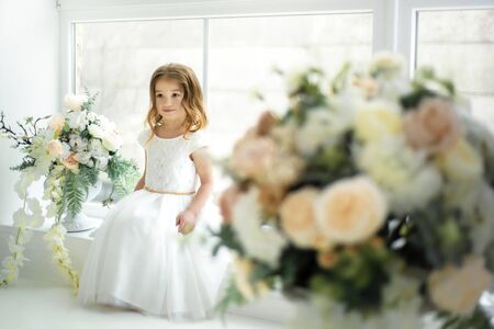 Little 3 years old beautiful tender smiling girl in long white dress sitting on window sill among bouquets of flowers. Spring concept. Child in flowers. Studio photoshoot. Indoorsの写真素材