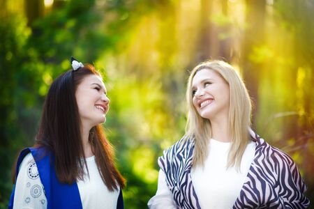 Portrait of two happy caucasian women, blonde and brunette, talking and smiling in sunset time in sunny park. Horizontal shape, copy space. Time together after selfisoltion. Friends meeting.の写真素材