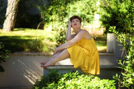 Young beautiful caucasian bored displeased girl with short haircut in short yellow dress sitting outdoors and thinking. sad barefoot short-haired woman in her garden in sunny day. Summer vacation 2020の写真素材