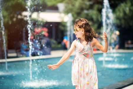 Loving life. Cheerful little girl in summer light dress and yellow sunglasses having fun playing with fountain water splashes. Summer vacation at sea. Happy child in summer hot day.の写真素材