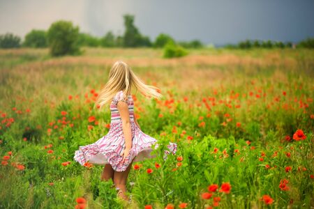 Faceless blonde girl dancing on meadow in poppies field in colorful dress before rain. Happy summer concept. Close to nature. Allergy time. Blonde hair and red poppy.の写真素材