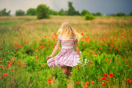 Faceless blonde girl walking on meadow in poppies field in colorful dress before rain. Happy summer concept. Close to nature. Allergy time. Blonde hair and red poppyの写真素材