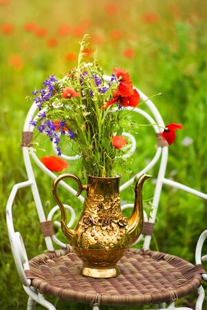 Vertical picture of bouquet from fresh wild flowers, chamomile and poppies, in golden teapot on chair in middle of meadow with poppies and green grass. Rural still life.の写真素材